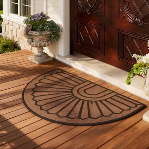 Può includere: Un zerbino marrone a forma di semicerchio con un design geometrico nero. Il tappeto è realizzato in un materiale resistente ed è posizionato su un portico in legno. Il design presenta un motivo a raggiera con linee curve.