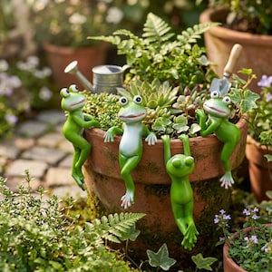 May include: Four green frog figurines are perched on the edge of a terracotta pot filled with greenery. The frogs have white bellies and are in various poses, adding a whimsical touch to the garden setting. A small watering can and trowel are visible.
