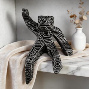 Puede incluir: Una escultura de metal gris oscuro de una persona con cortes geométricos. La figura tiene un brazo levantado y posa como si estuviera bailando. La escultura está sobre una superficie de mármol con un jarrón blanco y flores secas en el fondo.