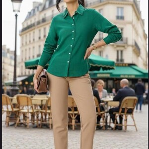 May include: A long-sleeved emerald green button-down shirt with a classic collar, paired with tan trousers that feature a straight-leg cut. The outfit is completed with brown heeled sandals and a small brown handbag held at the side.
