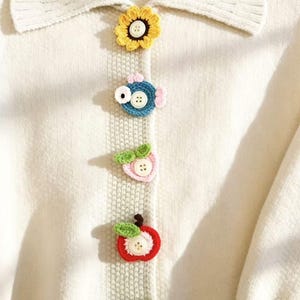 以下が含まれることがあります： リブ編みの襟と、4つの異なるかぎ針編みの装飾が施された縦型の前立てを備えたクリーム色のニットカーディガン。それぞれの装飾の中央には白いボタンが付いており、黄色いヒマワリ、青い魚、ピンクの桃、赤いリンゴがデザインされています。