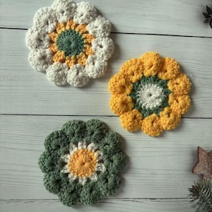 Pode incluir: Três bases de copos de flores de crochê feitas à mão em tons de branco, amarelo e verde. As bases de copos estão dispostas sobre uma superfície de madeira clara. Cada base de copo apresenta uma combinação de cores diferente, com um design floral central.