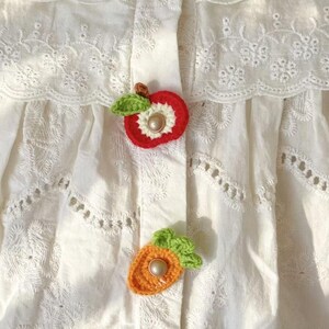 以下が含まれることがあります： 複雑な花柄のアイレット刺繍とスカラップエッジが特徴の白いコットンブラウス。フロントプラケットには、緑の葉が付いた赤いリンゴと、緑の葉が付いたオレンジ色のニンジンの手作りかぎ針編みアップリケが2つ取り付けられており、それぞれ中央にパールがあしらわれています。