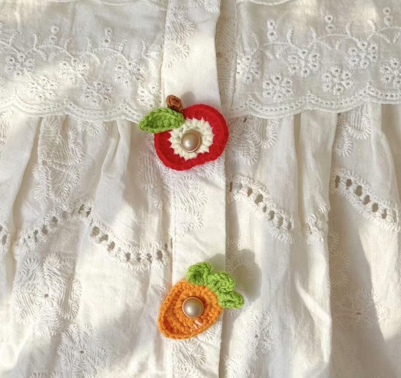 以下が含まれることがあります： 複雑な花柄のアイレット刺繍とスカラップエッジが特徴の白いコットンブラウス。フロントプラケットには、緑の葉が付いた赤いリンゴと、緑の葉が付いたオレンジ色のニンジンの手作りかぎ針編みアップリケが2つ取り付けられており、それぞれ中央にパールがあしらわれています。