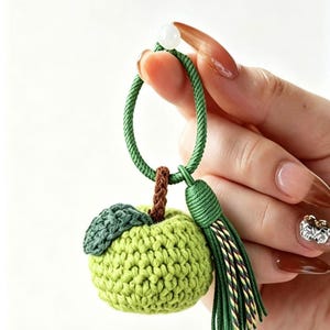 May include: A green crocheted apple keychain with a green loop, brown stem, and green leaf. A green tassel hangs from the loop. A white bead is at the top of the loop. The apple is a light green color.