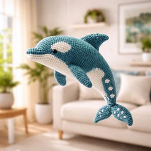 Puede incluir: Una orca tejida a mano en tonos azul y blanco, con ojos negros. El juguete hecho a mano presenta un diseño detallado, incluyendo aletas, cola y manchas blancas. La orca está suspendida en el aire, lo que sugiere que es una decoración colgante o un juguete.