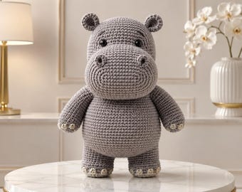 May include: A gray crocheted hippopotamus stuffed animal with a rounded body, small ears, and dark eyes. The hippo features a prominent snout and light-colored accents on the feet, standing upright with a textured yarn construction throughout.