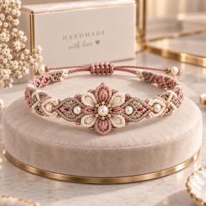 May include: A macrame bracelet featuring a central flower motif crafted from dusty pink and cream cord. The design incorporates small white pearls and gold-toned beads throughout the woven pattern. It includes an adjustable sliding knot closure.