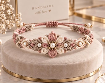 May include: A macrame bracelet featuring a central flower motif crafted from dusty pink and cream cord. The design incorporates small white pearls and gold-toned beads throughout the woven pattern. It includes an adjustable sliding knot closure.