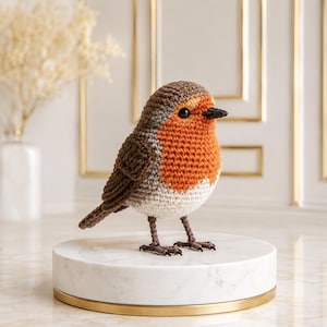 Può includere: Una statuetta di pettirosso all'uncinetto con testa e ali marroni, petto arancione brillante e ventre bianco. L'uccello ha un piccolo becco nero e occhi scuri, ed è posizionato su sottili zampe marroni con piedi dettagliati in filato.