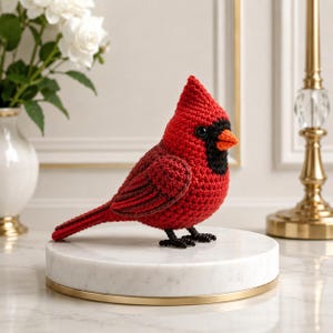 May include: A crocheted cardinal bird figurine featuring vibrant red yarn, a black mask around the eyes, a bright orange beak, and small black feet. The bird is crafted with a textured stitch pattern and has round, dark, glossy eyes.