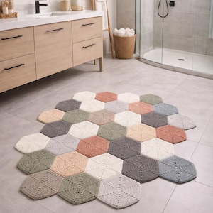 May include: A handmade, multi-colored crochet rug in a bathroom setting. The rug is composed of numerous hexagonal shapes in various pastel colors, including cream, gray, peach, and green. The rug is placed on a tiled floor, with a vanity and shower visible in the background.