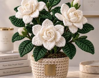 May include: A crocheted potted plant featuring three large white flowers and two buds with dark green leaves. The plant is housed in a beige crocheted pot with a small gold-colored rectangular plaque that reads Good Things Bloom Always.