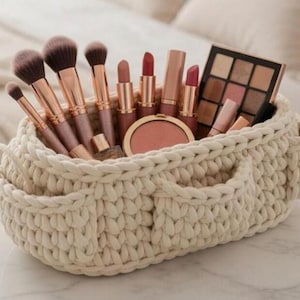 Puede incluir: Una cesta de ganchillo color crema llena de brochas de maquillaje, barras de labios, una polvera de colorete y una paleta de sombras de ojos. Las brochas tienen mangos de oro rosa y cerdas de color marrón oscuro. La cesta tiene dos pequeños bolsillos en la parte delantera.