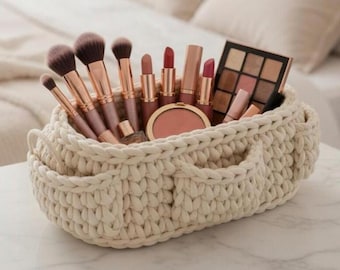 May include: A cream-colored, hand-crocheted basket filled with makeup brushes, lipsticks, a blush compact, and an eyeshadow palette. The brushes have rose gold handles and dark brown bristles. The basket has two small pockets on the front.