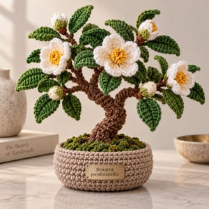 Peut inclure: Un bonsaï Stewartia pseudocamellia crocheté avec un tronc marron, des feuilles vertes et des fleurs blanches à cœur jaune. L'arbre est planté dans une base crochetée beige avec une plaque dorée portant l'inscription Stewartia pseudocamellia.
