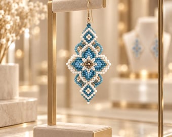 May include: A dangling earring featuring a geometric pattern crafted from small beads in shades of blue, white, and metallic gold. The earring has a symmetrical, elongated shape with a central flower-like motif and a gold-toned metal hook.