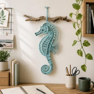 Könnte beinhalten: Ein hellblaues Makramee-Seepferdchen zum Aufhängen an der Wand, das an einem Stück Treibholz befestigt ist. Das Seepferdchen ist aufwendig gewebt mit einem detaillierten Design, das einen eingerollten Schwanz und einen strukturierten Körper aufweist. Das Seepferdchen ist etwa 50 cm hoch.