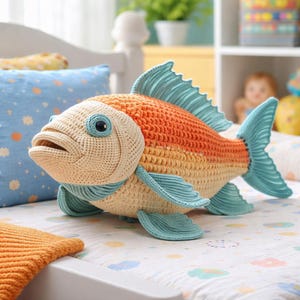 May include: A large, crocheted fish in shades of orange, yellow, and blue. The fish has large, round eyes and detailed fins. The plush toy is resting on a white surface with a patterned design.