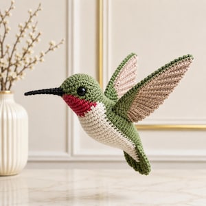 May include: A crocheted hummingbird figurine featuring a green head and back, a bright red throat patch, a cream-colored chest, and light brown wings with green edges. The bird has a long, thin black beak and a small, round black eye.