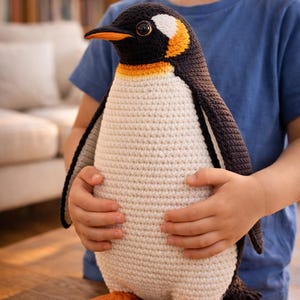 Peut inclure: Une peluche pingouin crochet&eacute;e &agrave; la main. Le pingouin a une t&ecirc;te, un dos et des ailes noirs, avec un ventre blanc. Il pr&eacute;sente des accents orange sur le cou et les pieds, et un bec noir. La peluche est tenue par une personne.