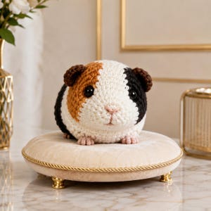 May include: A crocheted guinea pig plush toy featuring a tri-colour pattern of white, brown, and black yarn. The plush has dark round eyes, a small pink stitched nose, and brown ears, resting on a round, beige velvet cushion with gold trim.