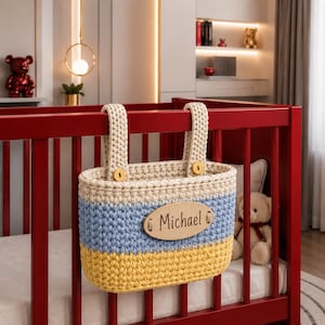 May include: A handmade crochet basket in cream, blue, and yellow hangs from a red crib. The basket has a wooden name tag that reads "Michael". The basket is secured with cream-colored straps and wooden buttons. A teddy bear is visible in the background.