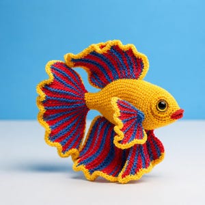 Puede incluir: Un pez Betta hecho a mano con ganchillo en colores vibrantes. El pez presenta un cuerpo amarillo, aletas a rayas rojas y azules y ribetes amarillos. El pez tiene un ojo detallado y labios rojos, sobre un fondo azul.