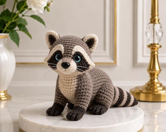 May include: A crocheted raccoon toy featuring a grey and cream-colored body, dark brown paws, and a striped tail. The raccoon has large, bright blue eyes, a black nose, and thin whiskers, crafted with a detailed yarn texture throughout its form.