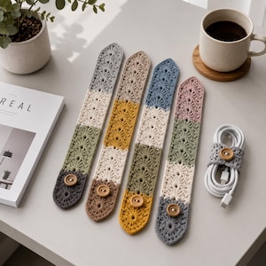 Könnte beinhalten: Vier handgefertigte Kabelorganisatoren aus Häkelarbeit in verschiedenen Farben: Grau, Olivgrün, Beige, Gelb, Blau und Rosa. Jeder Organizer hat einen Knopfverschluss. Ein fünfter Organizer umwickelt ein weißes Ladekabel. Eine Kaffeetasse und ein Magazin sind ebenfalls im Bild.