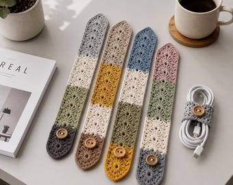 May include: Four handmade crochet cable organizers in various colors: gray, olive green, tan, yellow, blue, and pink. Each organizer features a button closure. A fifth organizer wraps a white charging cable. A cup of coffee and a magazine are also in the image.
