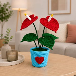 Puede incluir: Una planta de Anthurium hecha a mano con dos flores rojas vibrantes, hojas verdes y una maceta azul adornada con un corazón rojo. Las flores tienen detalles blancos y amarillos. Es un artículo decorativo.