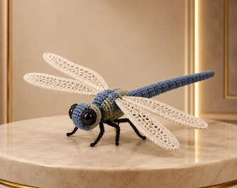 May include: A crocheted dragonfly toy featuring a blue and light green segmented body, four intricate cream-colored lace-like wings, and large black glossy eyes. The insect has six thin black legs and a long, slender, textured blue tail.