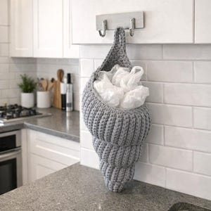 Op de afbeelding: Een grijze, gehaakte schelpvormige tas die aan een muur hangt. De tas is gevuld met witte plastic zakken. De tas is opgehangen aan een metalen haak. Het aanrecht en de witte tegelmuur zijn op de achtergrond.