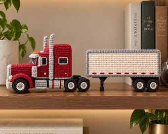 May include: A crocheted semi-truck toy featuring a red cab with grey accents and a long, rectangular beige trailer with grey trim. The truck has black wheels with white centers, two tall grey exhaust stacks, and small red lights along the trailer side.