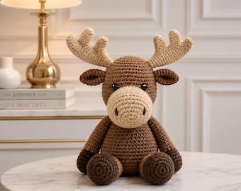 May include: A brown crocheted moose stuffed animal with a cream-colored muzzle and large cream-colored antlers. The toy features dark brown limbs, a round body, and two black plastic eyes. It is seated upright with its arms resting at its sides.