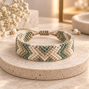 May include: A woven macrame bracelet featuring a geometric chevron pattern in sage green, cream, and tan thread. The bracelet includes an adjustable sliding knot closure with two small, round, light-colored wooden beads at the ends of the cords.