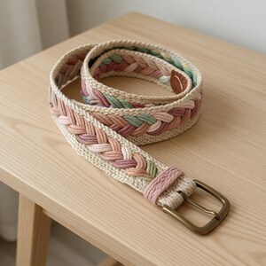 May include: Braided belt with a brass buckle. The belt has a woven design with sections of pink, green, and beige. The belt is on a light-coloured wooden surface.