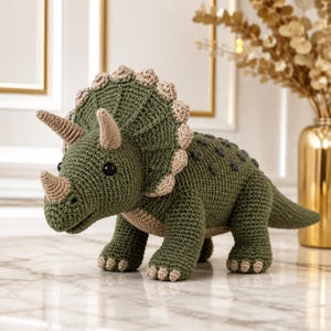 Puede incluir: Un juguete de dinosaurio triceratops tejido a ganchillo en color verde oliva con una gola, cuernos y vientre en color beige. El dinosaurio presenta ojos redondos oscuros, una espalda texturizada con pequeños bultos verde oscuro y cuatro patas robustas.