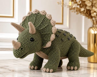 May include: A crocheted triceratops dinosaur toy in olive green with a beige frill, horns, and underbelly. The dinosaur features dark, round eyes, a textured back with small dark green bumps, and four sturdy legs with beige-colored claw details.
