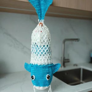 May include: A handmade crochet whale-shaped bag in shades of blue and white. The bag features a blue head with black and white eyes, and a white net-like body. The whale is hanging from a wooden cabinet.