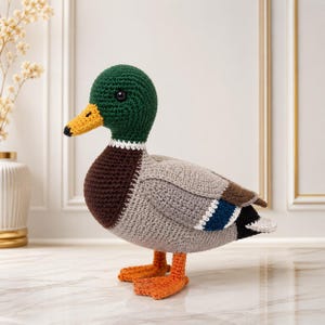 May include: A crocheted mallard duck toy featuring a dark green head, a yellow beak, a dark brown chest, and a light gray body with blue and white wing accents. The duck has orange webbed feet and black bead eyes, crafted with yarn.