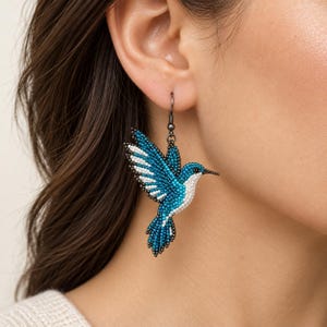 May include: A hummingbird-shaped earring crafted from tiny beads in shades of teal, blue, and white. The bird is depicted in a mid-flight position with wings extended, suspended from a dark metal hook that attaches to the earlobe.