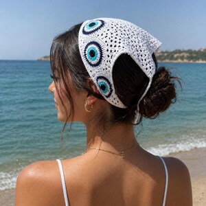 Puede incluir: Pañuelo blanco de ganchillo con tres diseños de ojo turco azul y negro. El pañuelo se lleva en la cabeza y se ata en la parte posterior. El fondo es una playa con agua azul y un cielo despejado.
