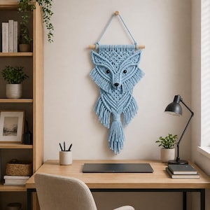May include: A light blue macrame fox wall hanging. The fox has a detailed design with fringe and a wooden dowel for hanging. The fox's eyes and nose are black. The wall hanging is a decorative piece.