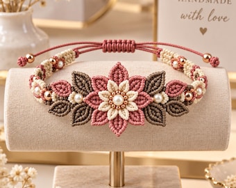 May include: A macrame bracelet featuring a central flower motif in cream and dusty rose, flanked by dark brown leaf-shaped knots. The band includes small white pearls and metallic gold beads, finished with a sliding knot closure and gold-toned end beads.