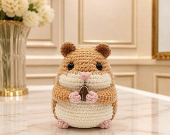 May include: A small, round, crocheted hamster toy made with tan and cream-colored yarn. The hamster has black plastic eyes, a small pink stitched nose, and tiny pink paws holding a single dark brown sunflower seed in its front.