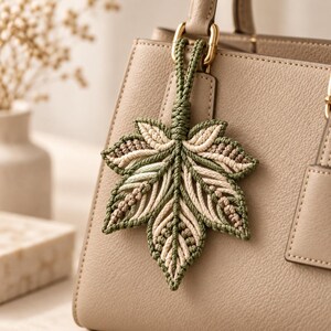 Può includere: Un portachiavi a forma di foglia in macramè nei toni del verde salvia e crema, caratterizzato da intricati motivi a cordoncino annodato. La foglia è fissata a un anello in metallo dorato e pende dal manico di una borsa in pelle beige.