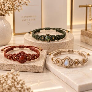 May include: Three macrame bracelets featuring central gemstones. One is dark green with a round stone, one is rust-colored with a round stone, and one is cream-colored with a teardrop-shaped stone. Each bracelet includes small gold-toned metal beads.