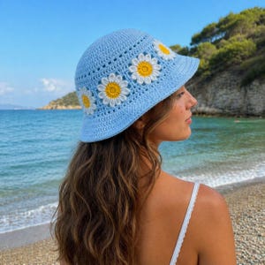 Peut inclure: Un chapeau de pêcheur en crochet bleu clair avec des appliqués de marguerites blanches et jaunes. Le chapeau a un large bord. L'arrière-plan montre une plage et l'océan.